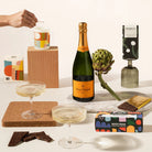 A styled gift set featuring a bottle of Veuve Clicquot champagne, a box of Rocky Road chocolate, a candle, and a pair of Ferm Living champagne glasses.