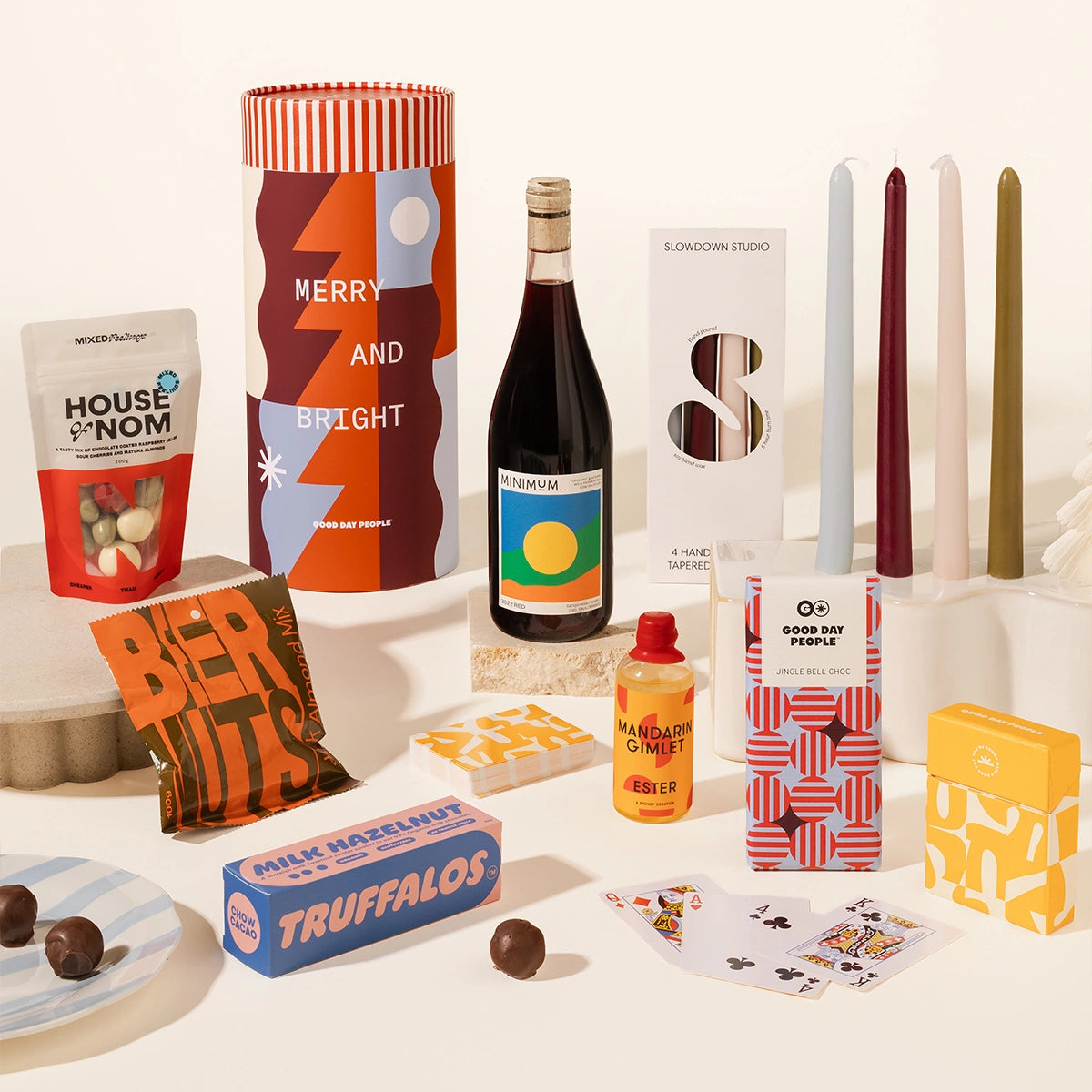 A curated gift assortment arranged on a white surface against a warm off-white background, including a tall cylindrical box with red, orange, and blue geometric artwork reading 'MERRY AND BRIGHT,' a dark bottle with a colorful label, several candles in white, burgundy, pale blue, cream, and olive tones, a bag of House of Nom snacks, a bag of Beer Nuts, a blue box of Truffalos, a small orange bottle, patterned boxes, playing cards, and chocolate pieces.
