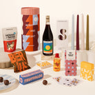 A curated gift assortment arranged on a white surface against a warm off-white background, including a tall cylindrical box with red, orange, and blue geometric artwork reading 'MERRY AND BRIGHT,' a dark bottle with a colorful label, several candles in white, burgundy, pale blue, cream, and olive tones, a bag of House of Nom snacks, a bag of Beer Nuts, a blue box of Truffalos, a small orange bottle, patterned boxes, playing cards, and chocolate pieces.