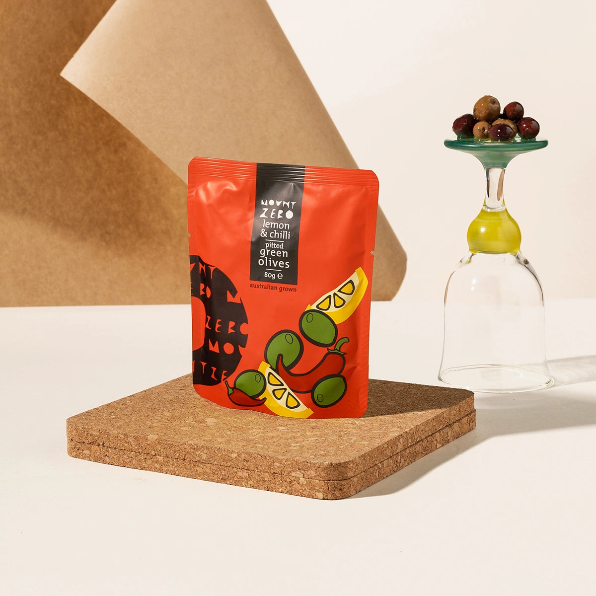A pouch of Mount Zero Lemon & Chilli Green Olives sits on a cork coaster next to a clear stemmed glass with a green base holding a small pile of olives, against a warm beige studio background with a brown paper prop.