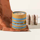 Olsson's Sea Salt Flakes Eyre Peninsula pure sea salt flakes in a kraft paper canister with blue stripes, styled with brown paper and sliced salt bars on a white surface.