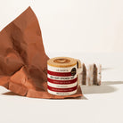 A jar of Olsson's Red Gum Smoked Salt sits on a white surface next to a crumpled brown paper bag and two salt blocks, photographed in a clean studio setting.