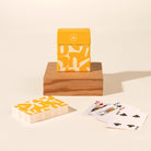 A deck of patterned playing cards with a matching yellow and white box sits on a wooden block, with a second deck and a few fanned cards on a cream background.