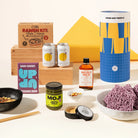 A curated flat-lay of multiple packaged food and beverage products on a warm cream background, including a ramen kit box, two cans, a bottle of Mildly Wild chilli oil, a jar of Moji matcha spread, a bowl of ramen noodles, chopsticks, and a black dish. Ramen Riko Japanese inspired gourmet gift hamper