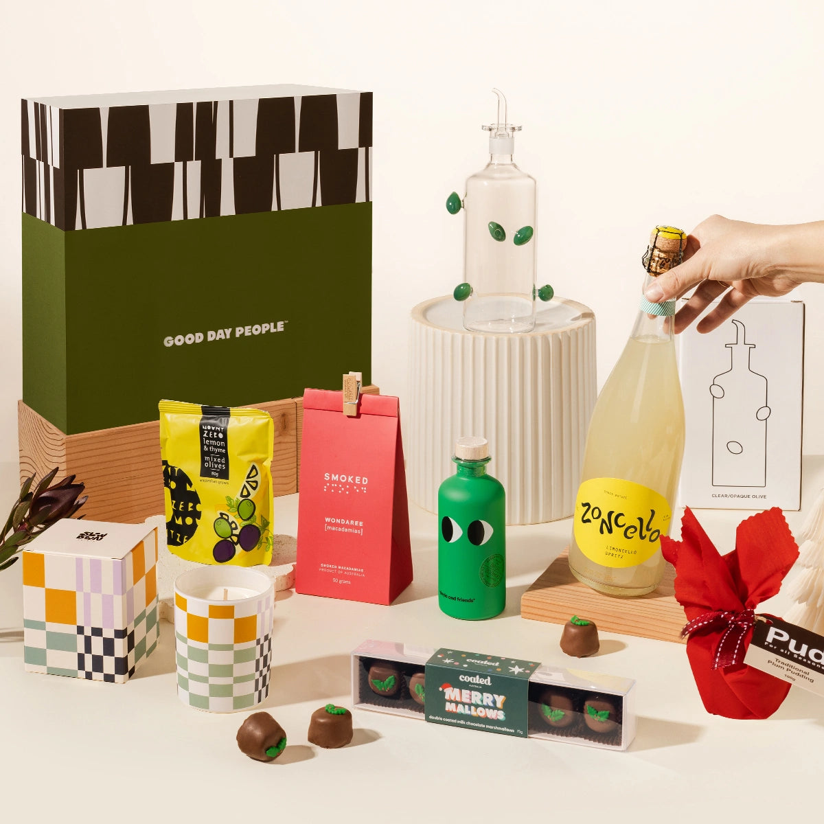 A curated gift set arrangement on a white surface featuring a large olive-green and black patterned box labeled GOOD DAY PEOPLE, a hand holding a  yellow limoncello spritz bottle with a yellow label, an olive oil holder bottle from maison balzac, macadamias, olives, olive oil from yia yia, a red mini christmas pudding, a slowdown studio candle and chocolate coated marshmallows