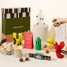 A curated gift set arrangement on a white surface featuring a large olive-green and black patterned box labeled GOOD DAY PEOPLE, a hand holding a  yellow limoncello spritz bottle with a yellow label, an olive oil holder bottle from maison balzac, macadamias, olives, olive oil from yia yia, a red mini christmas pudding, a slowdown studio candle and chocolate coated marshmallows