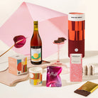 A styled gift hamper featuring a bottle of wine, a cylinder labeled GOOD DAY PEOPLE, a candle, fairy floss, a chocolate bar, with a large pink abstract backdrop behind them.