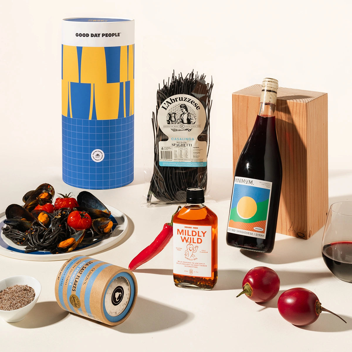 Salty Simon Gift Hamper from Good Day People. A styled food and drink assortment on a light beige background, including mussels with tomatoes, a bottle of red wine, a bottle of amber liquor, packaged black pasta, a blue and yellow cylindrical container, a small spice jar, a red chili pepper, and a glass of red wine.