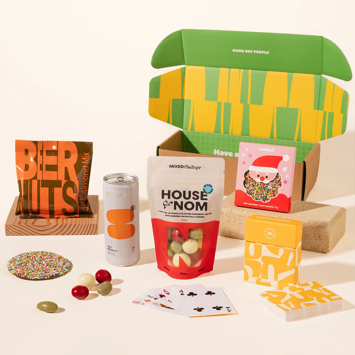 A curated gift box set arranged on a light beige studio background. The set includes a green and yellow patterned box, a bag of mixed nuts, a can of soda, a bag of House of Nom choc coated nuts, a speckled milk choc, a yellow playing card deck.