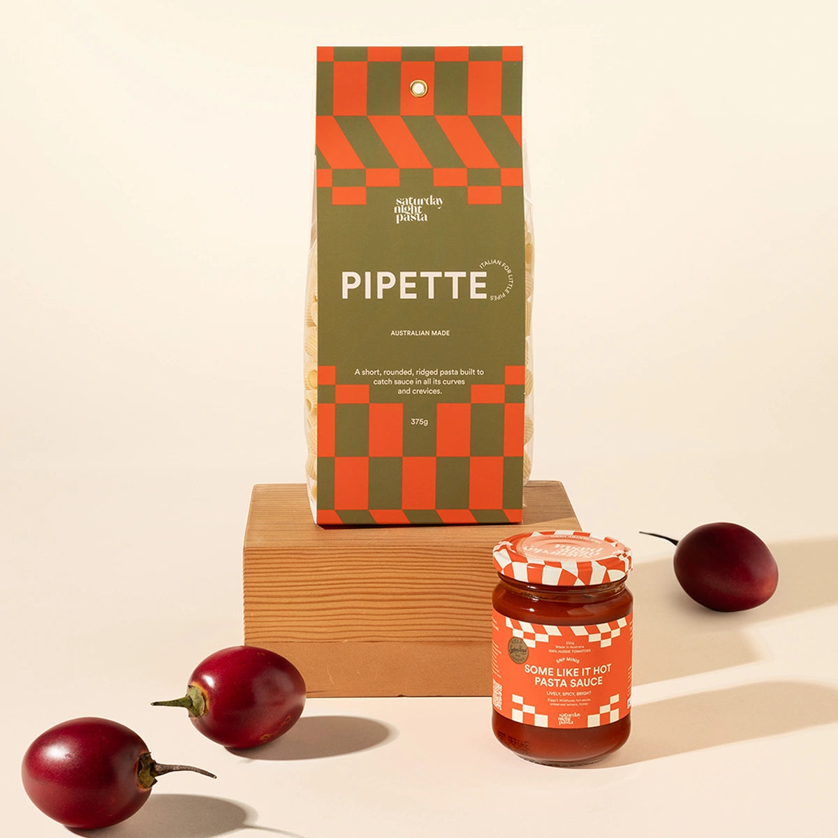 A styled product shot featuring a bag of Pipette Australian Made pasta and a jar of pasta sauce on a wooden block, with red chili peppers scattered around on a warm cream background.