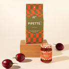 A styled product shot featuring a bag of Pipette Australian Made pasta and a jar of pasta sauce on a wooden block, with red chili peppers scattered around on a warm cream background.