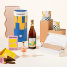 A curated gift set featuring a blue and yellow cylindrer labeled Good Day People, a bottle of rosé wine, a pink salted caramel milk chocolate bar, a small pink bottle containing lychee martini, a blush sleep mask in an open box, and colorful candles and wooden blocks on a clean cream background.
