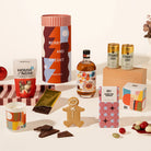 A curated gift set arranged on a light neutral surface, featuring a tall cylindrical box with red-and-white stripes and the words “MERRY AND BRIGHT,” a bottle of liquor, two small cans of tonic, a bag of chocolates from House of Nom, a gingerbread cookie, chocolate pieces, a candle, a patterned box, and other matching gift items.