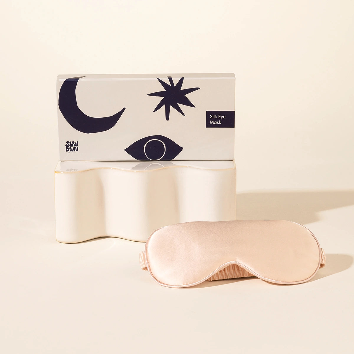 An artsy navy and white packaging box sits on a wavy pedestal. Next to it, a silk eye mask in a nude colour is displayed.