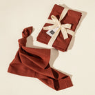 A rust-colored linen napkin set, including a folded cloth tied with a cream ribbon and a loose matching cloth, displayed on a warm off-white background.