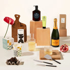 A gift set on a warm off-white background, featuring a bottle of Veuve Champagne, Leif products, a wooden serving board, a Ferm living carafe, chocolate and cheese knives.
