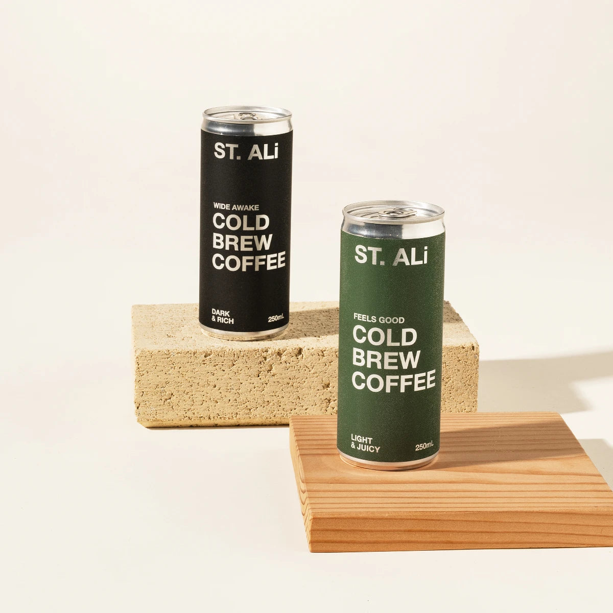 Two ST. ALI Cold Brew Coffee cans, one black and one dark green, displayed on stacked wooden blocks against a warm off-white background.