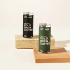 Two ST. ALI Cold Brew Coffee cans, one black and one dark green, displayed on stacked wooden blocks against a warm off-white background.
