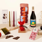 Gift hamper assortment with a bottle of wine, tapered candles (set of 4), a mini christmas pudding wrapepd in red fabric, a block of chocolate in a christmassy red and blue wrapper. Cylindrical packaging with a festive, fun design.