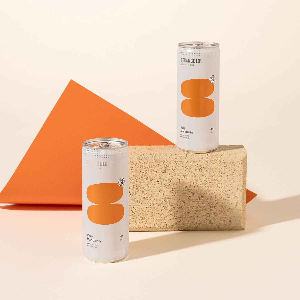 Two strangelove mandarin soda cans styled on and in front of a beige stone block with an orange geometric sheet behind them on a warm cream background.