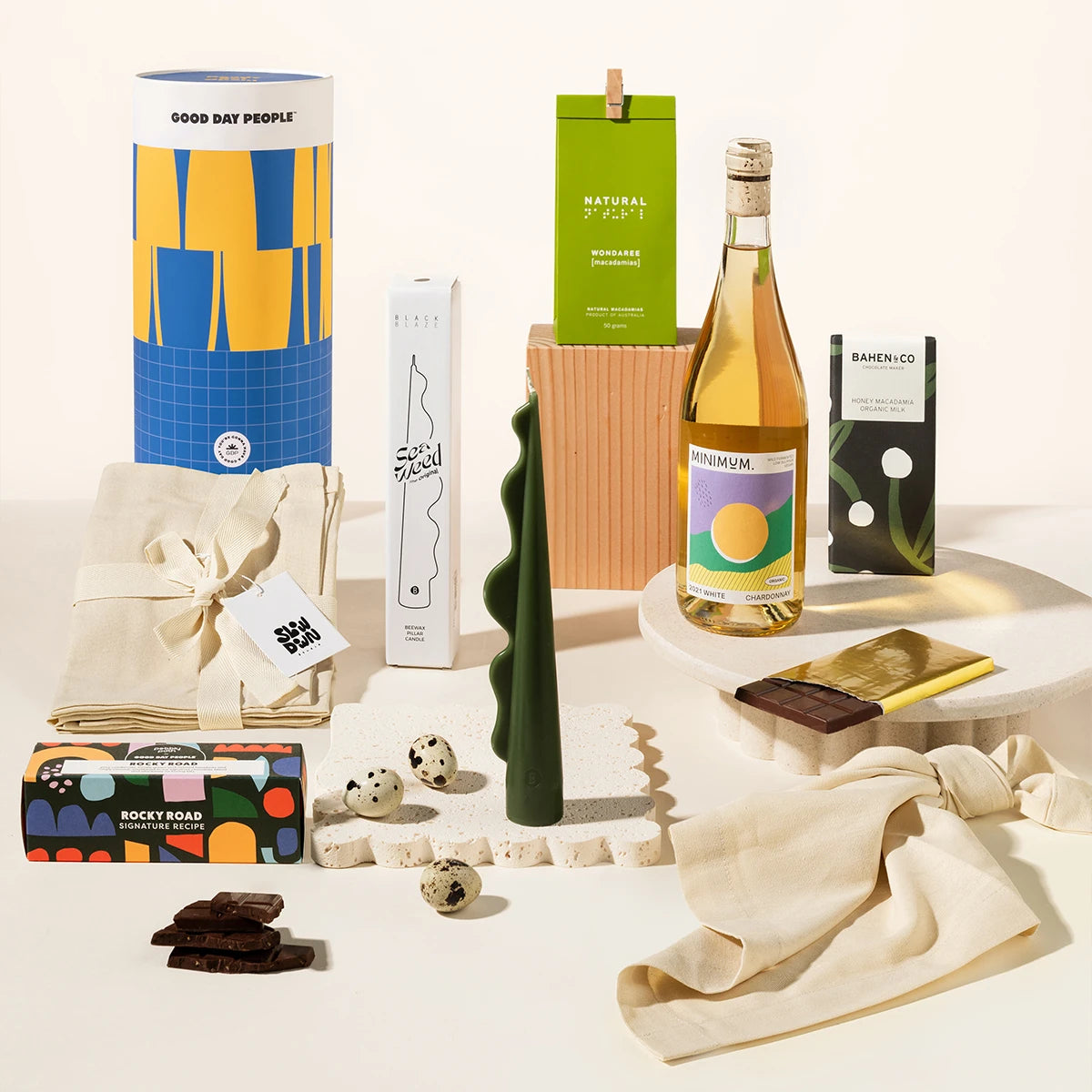 A curated gift assortment on a clean white studio background, including a blue and yellow cylindrical container, a green box labeled of macadamias, a bottle of wine, a dark green sculptural candle, chocolate, linen napkins and rocky road.