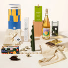 A curated gift assortment on a clean white studio background, including a blue and yellow cylindrical container, a green box labeled of macadamias, a bottle of wine, a dark green sculptural candle, chocolate, linen napkins and rocky road.