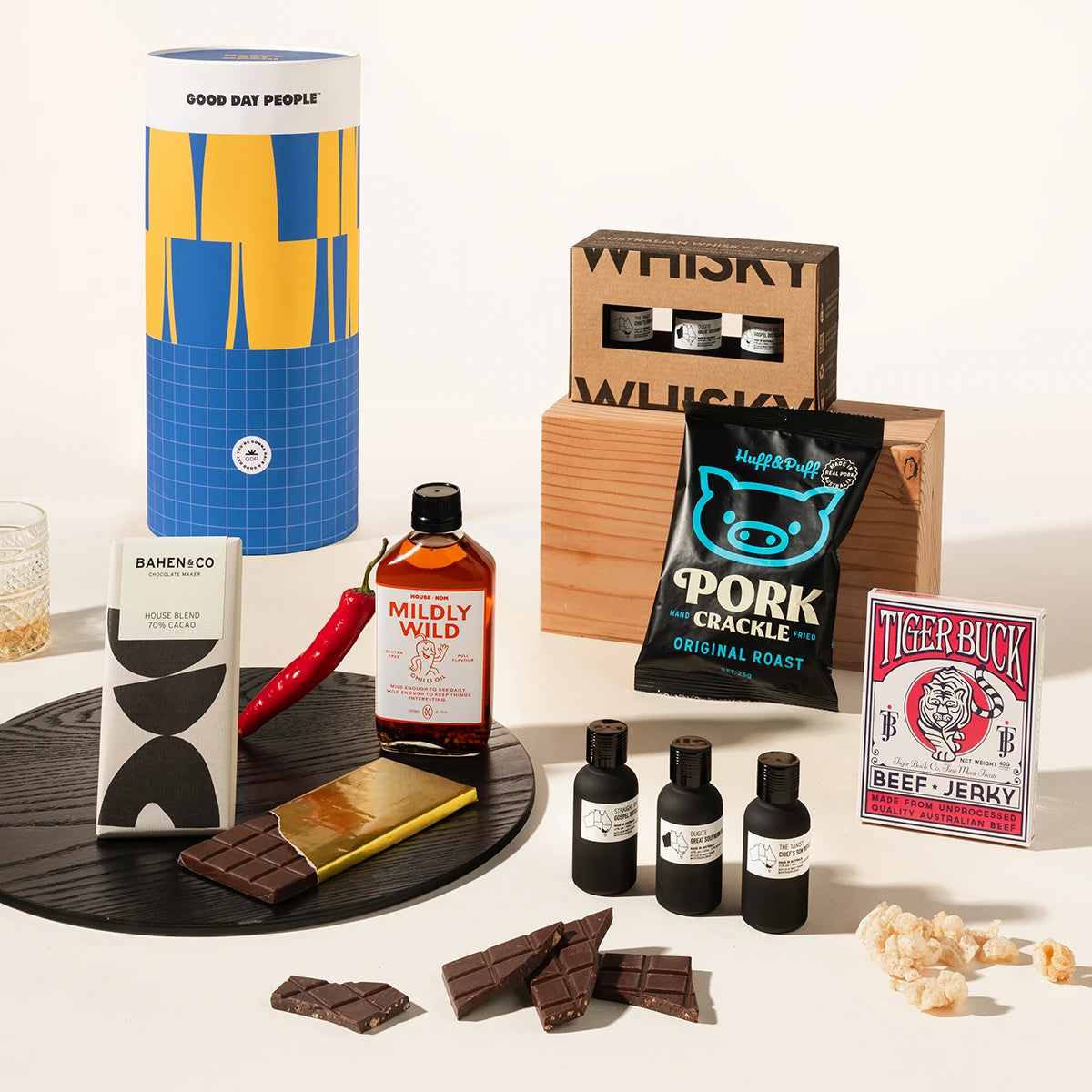 A curated gift set arranged on a white surface with a black round tray. Visible items include a tall blue-and-yellow cylindrical package labeled Good Day People, a wooden box with whiskey bottles, a bag of Pork Cracker beef jerky, a chocolate bar, a Mildly Wild chilli oil bottle, three small dark bottles, chocolate pieces, a red chili pepper, a gold chocolate piece, and a Tiger Buck beef jerky box.