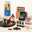 A curated gift set arranged on a white surface with a black round tray. Visible items include a tall blue-and-yellow cylindrical package labeled Good Day People, a wooden box with whiskey bottles, a bag of Pork Cracker beef jerky, a chocolate bar, a Mildly Wild chilli oil bottle, three small dark bottles, chocolate pieces, a red chili pepper, a gold chocolate piece, and a Tiger Buck beef jerky box.