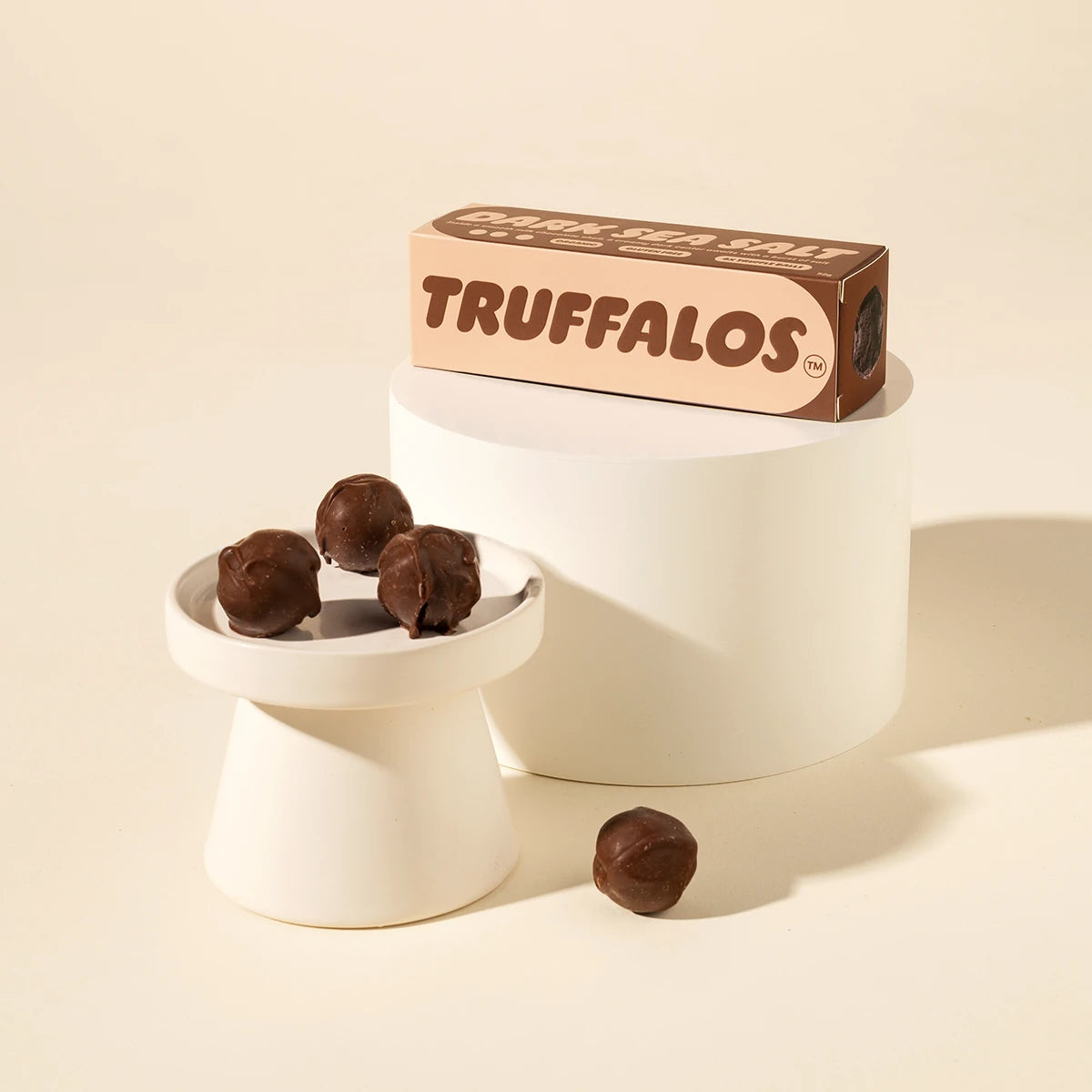 A box of Truffalos chocolate truffles displayed on a white pedestal, with three chocolate truffles on a small white dish and one loose in front, against a warm cream background.