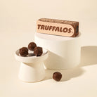 A box of Truffalos chocolate truffles displayed on a white pedestal, with three chocolate truffles on a small white dish and one loose in front, against a warm cream background.