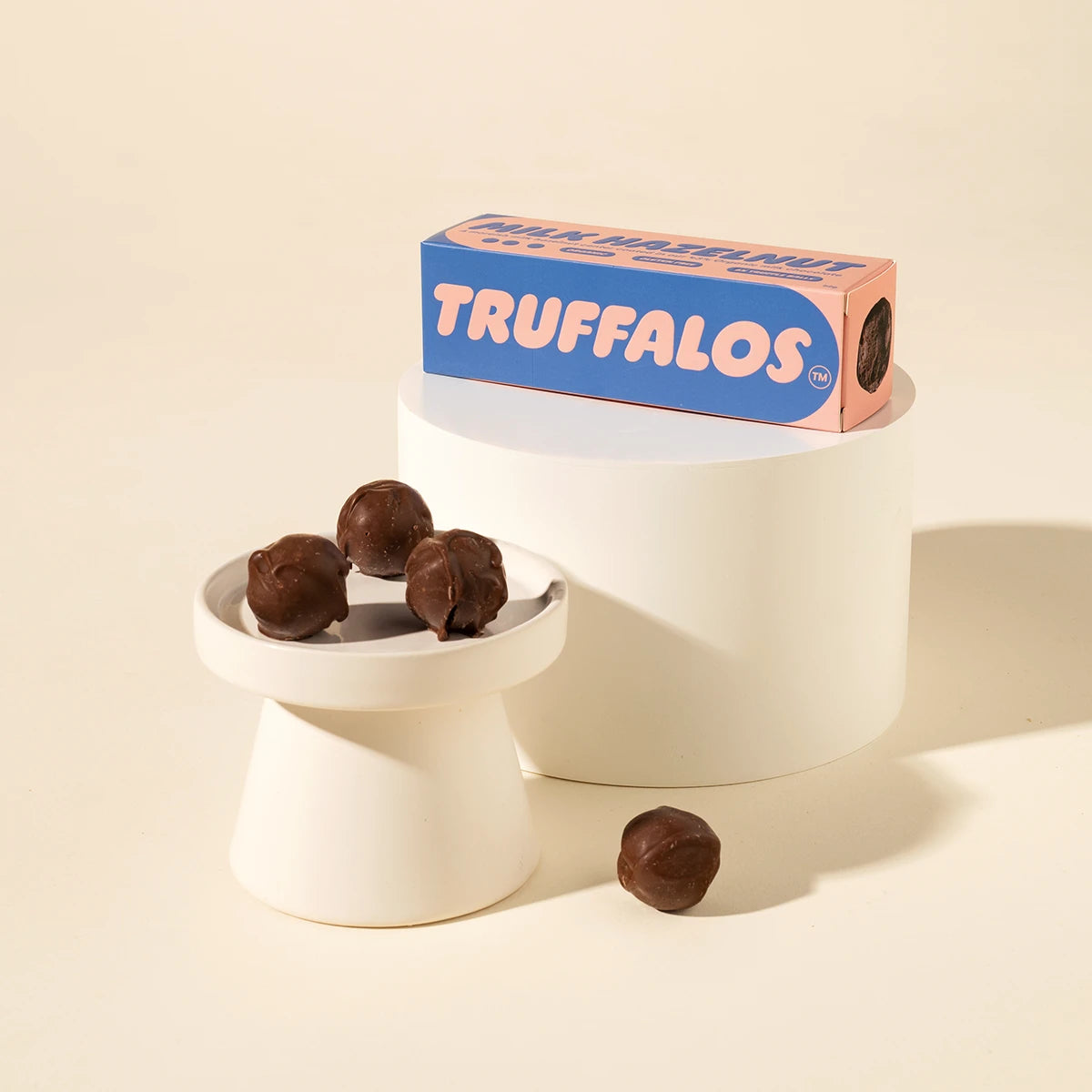 A box of Truffalos chocolate truffles displayed on a white pedestal next to a small dish holding three chocolate truffles, with one loose truffle on the surface, set against a warm cream background.