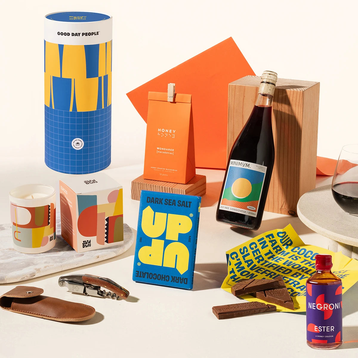 A curated gift set featuring a blue and yellow cylinder, macadamias, a bottle of Negroni, a candle, chocolates, a corkscrew, and a bottle of wine.