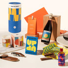 A curated gift set featuring a blue and yellow cylinder, macadamias, a bottle of Negroni, a candle, chocolates, a corkscrew, and a bottle of wine.