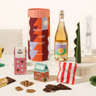 A curated gift set featuring a Christmassy designed canister as the packaging, a bottle of whitewine; a green wavy decorative candle, a block of chocolate with a geometric red and blue design labelled Jingle Bell Choc, a gingerbread house shaped box with pretzels in it and a red and white striped pouch containing fairy floss. 